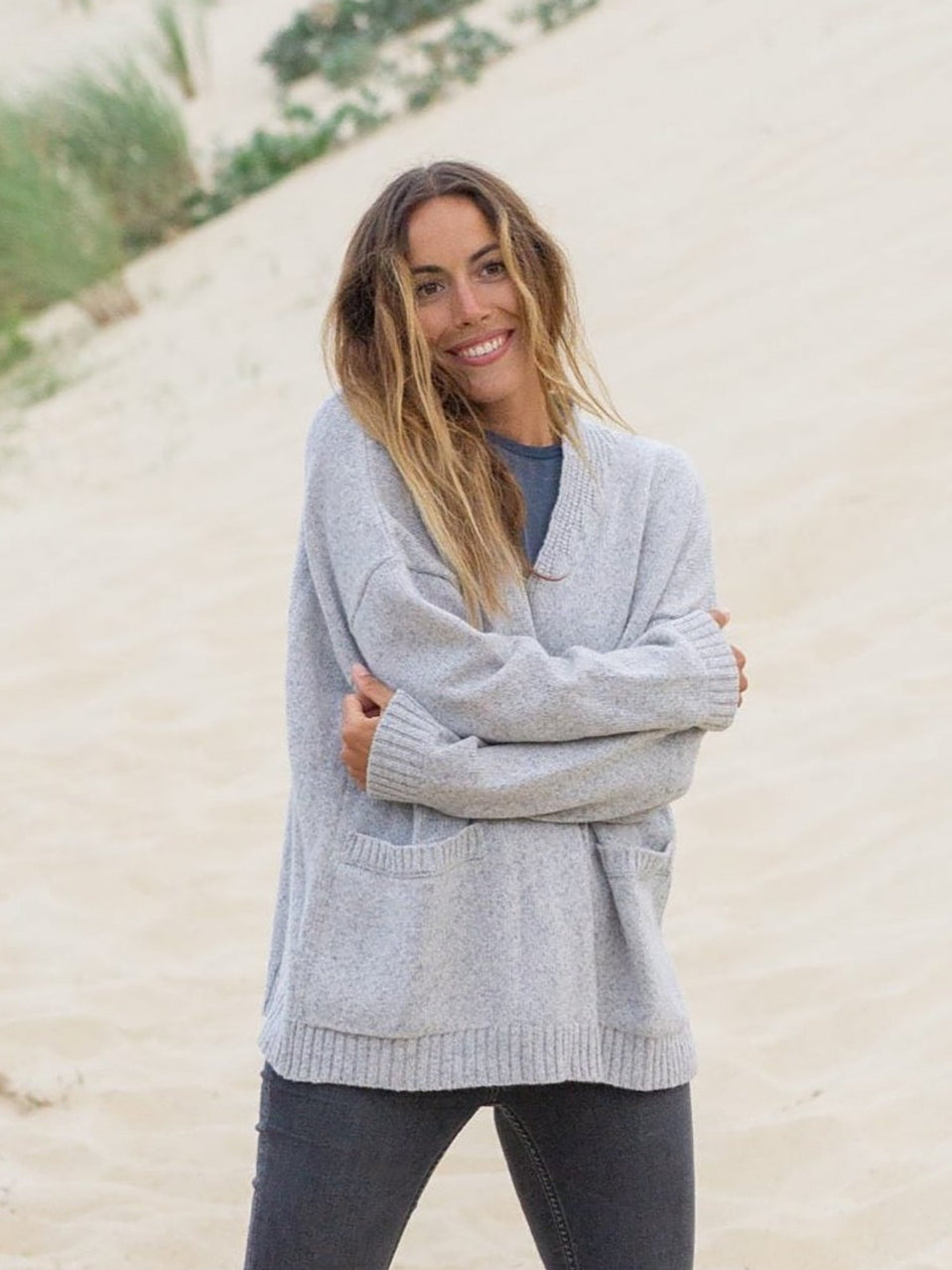 Graumelierter Oversized Frauen Cardigan aus 100% Merino Schurwolle von FUXBAU. Getragen von einem braunhaarigem Model am Strand in Fankreich.