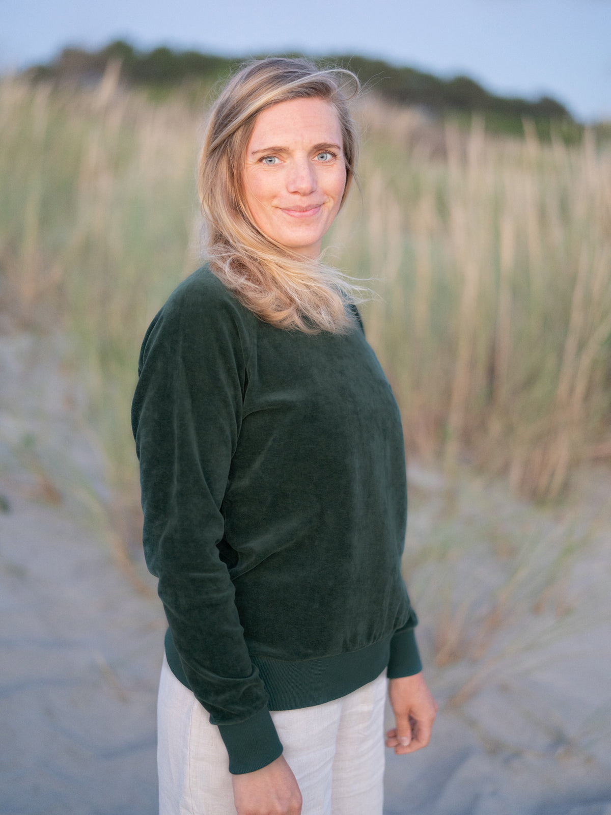 Isi trägt den FUXBAU Frauen Samt Sweater in Grün in den Dünen in Portugal – 100 % Bio-Baumwolle, GOTS-zertifiziert, Made in Portugal