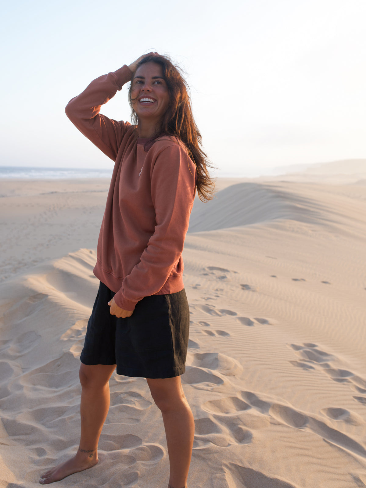 Frau trĂ€gt stĂŒckgefĂ€rbten roten FUXBAU Pullover aus Biobaumwolle am Strand in Portugal