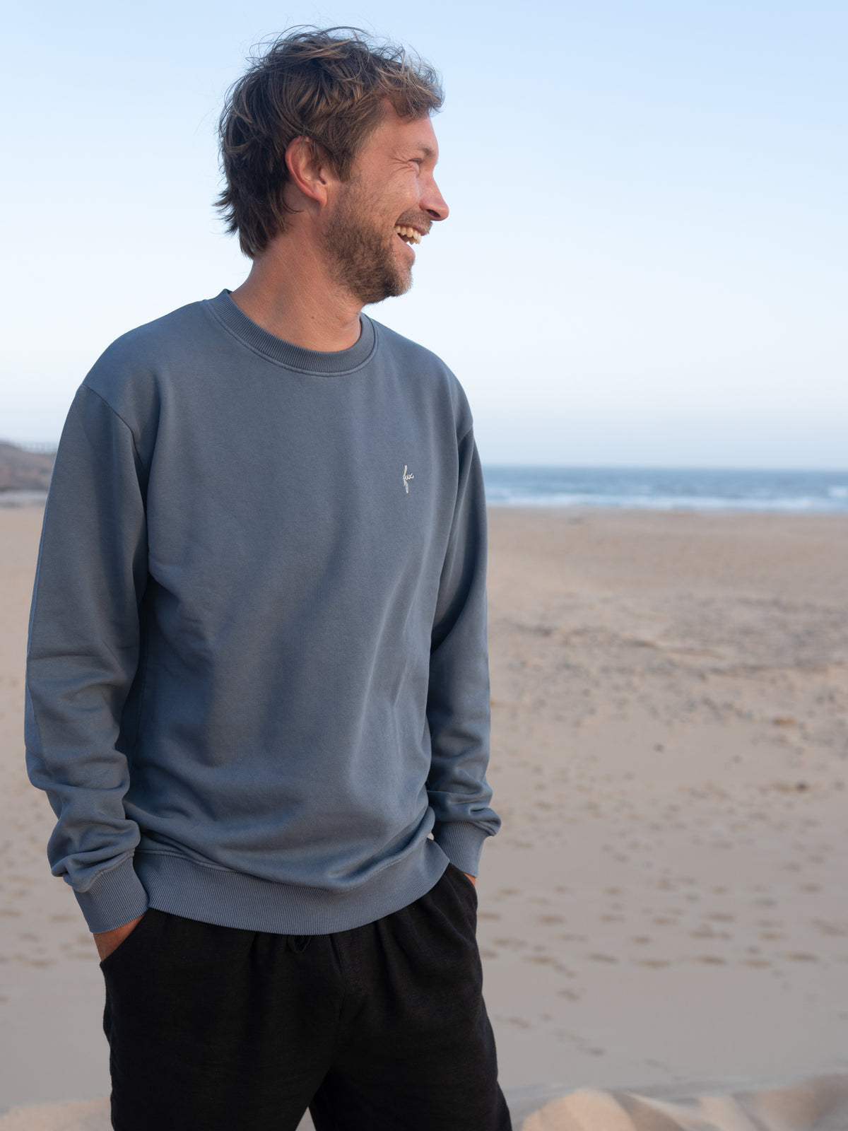 Mann trÀgt fair gefertigten seeblauen FUXBAU Pullover aus Biobaumwolle vor Strand und Meer