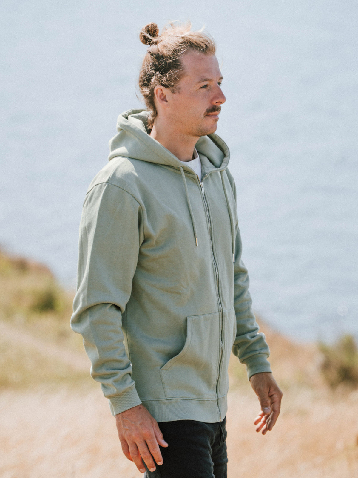 Unisex Sweatjacke in Seegras aus Biobaumwolle getragen von einem Mann an der Küste