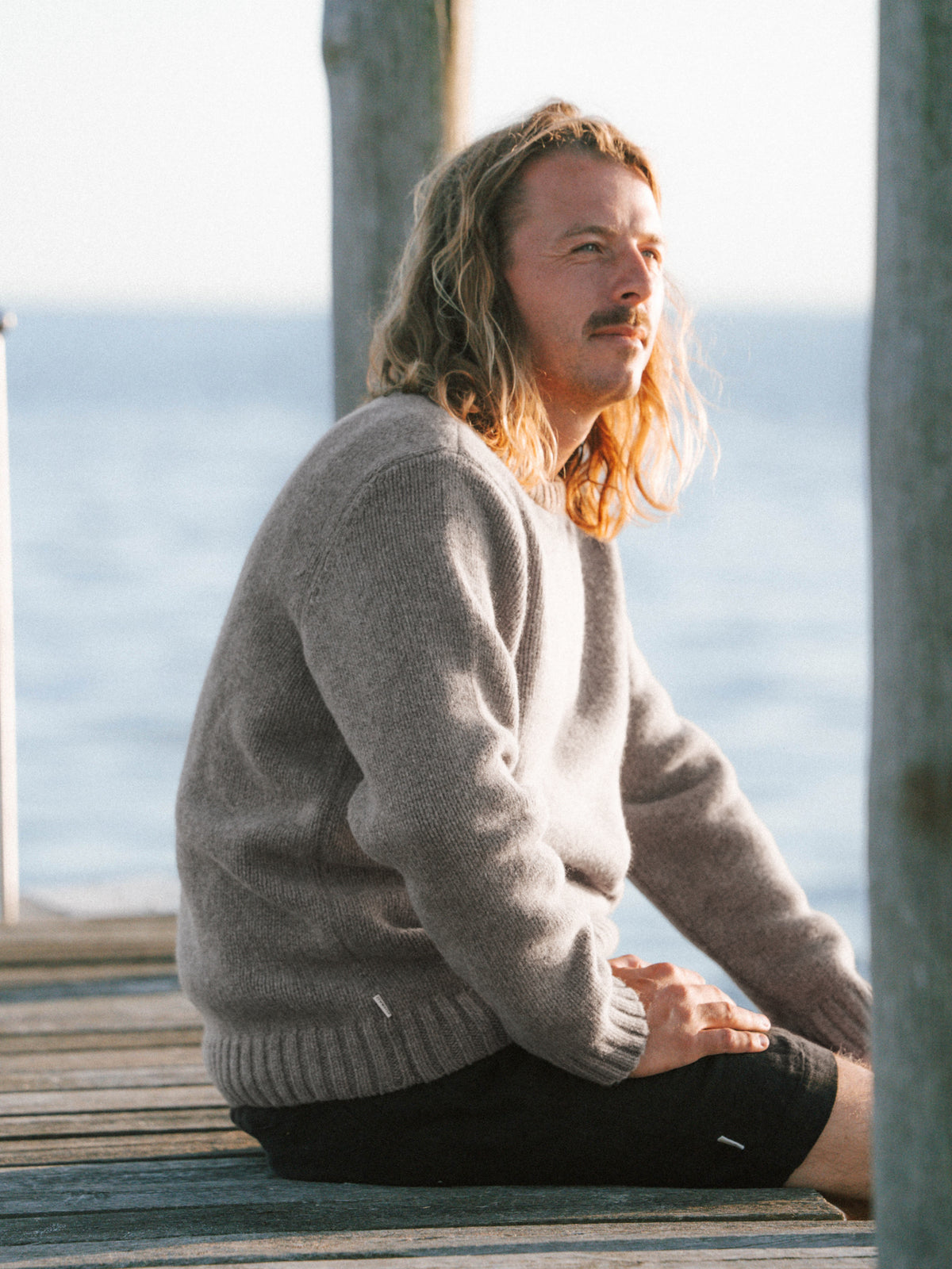 Ole sitzt auf einem Steg am Meer und trÀgt den MÀnner Strickpullover in braunmeliert aus 100% Merinowolle der in Deutschland hergestellt wird.