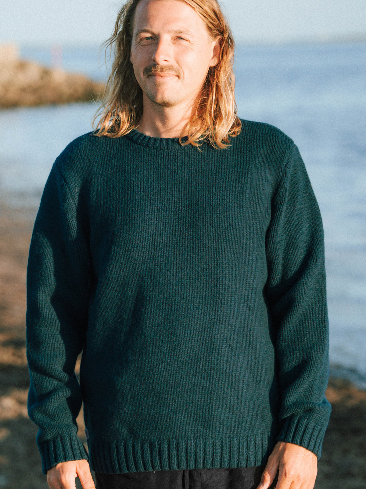 Ole trägt am Meer den Männer Merino Strickpullover in grün der in Deutschland hergestellt wird - Gesamtansicht
