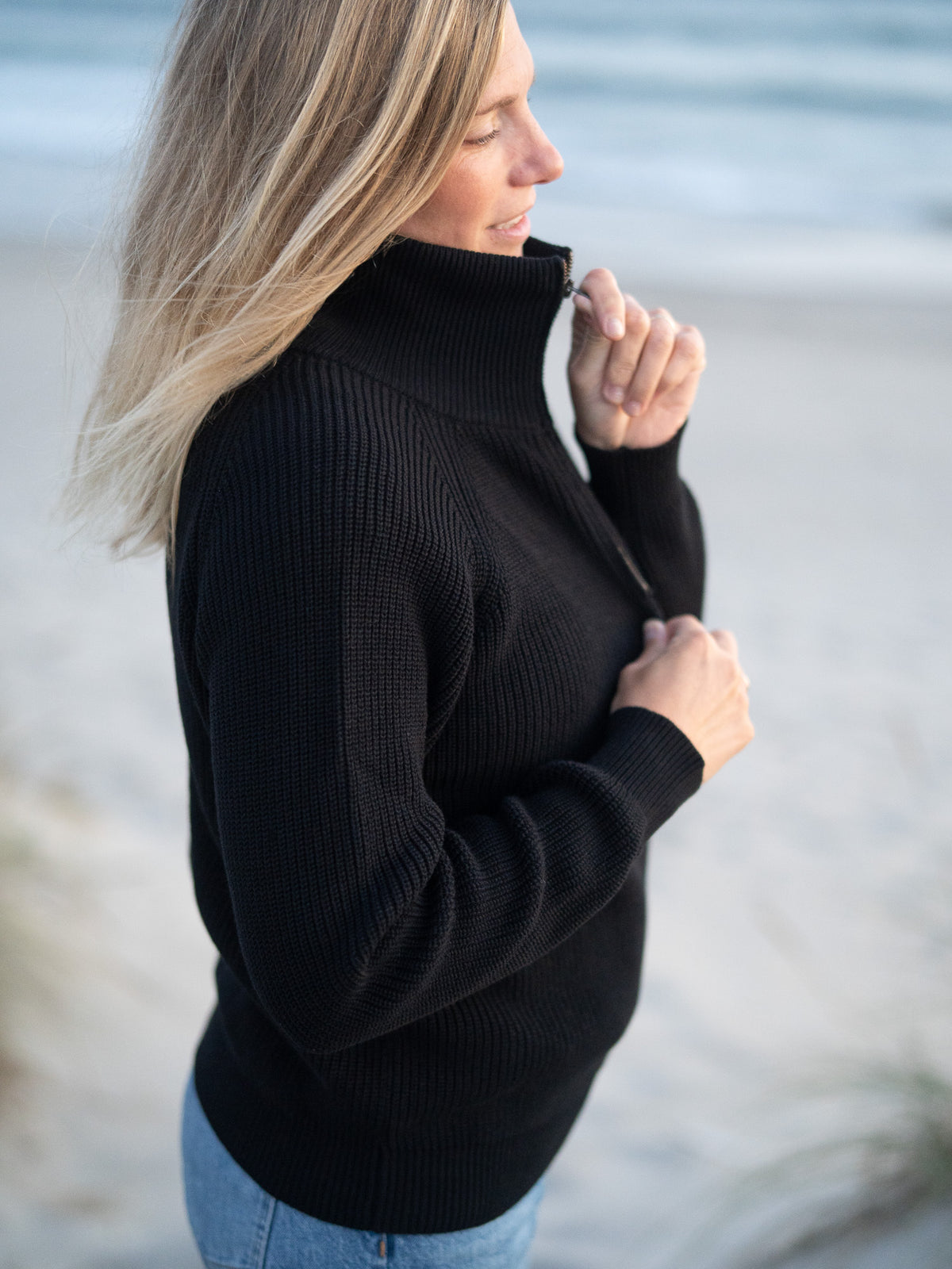 Eine junge, blonde Frau trägt einen Fair Fashion 4Seasons Troyer aus Biobaumwolle Made in Italy vom Fair Fashion Label FUXBAU am Strand in Portugal. Seitliche Ansicht.