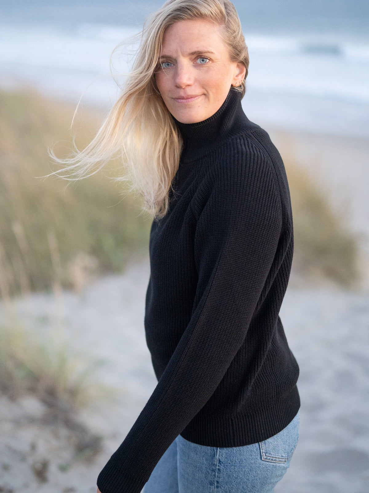 Eine junge, blonde Frau trägt einen Fair Fashion 4Seasons Troyer aus Biobaumwolle Made in Italy vom Fair Fashion Label FUXBAU am Strand in Portugal. Seitliche Ansicht.