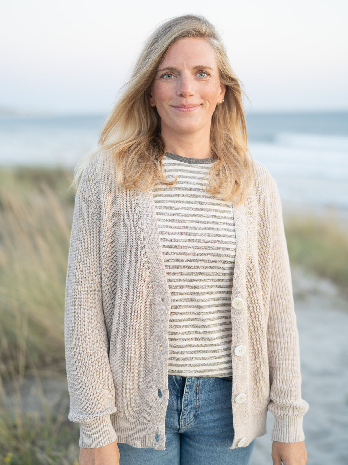 Eine junge, blonde Frau trÀgt eine beige melierte 4Seasons Strickjacke vom Fair Fashion Modelabel FUXBAU aus 100% GOTS Biobaumwolle am Strand in Portugal.