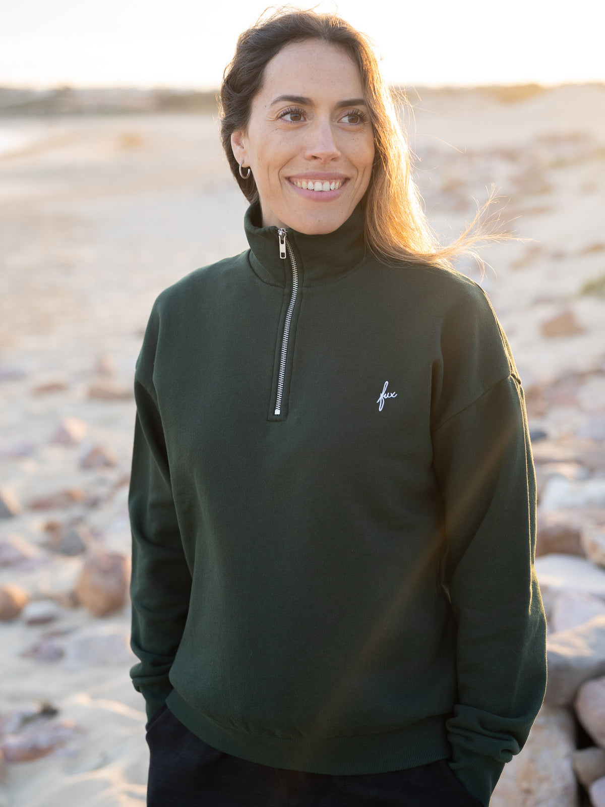 Junge, braunhaarige Frau trĂ€gt einen Fair Fashion Frauen Half Zip Sweater von FUXBAU in grĂŒn aus gots zertifizierter Biobaumwolle Made in Portugal mit YKK ReisverschlĂŒssen am Strand in Portugal.