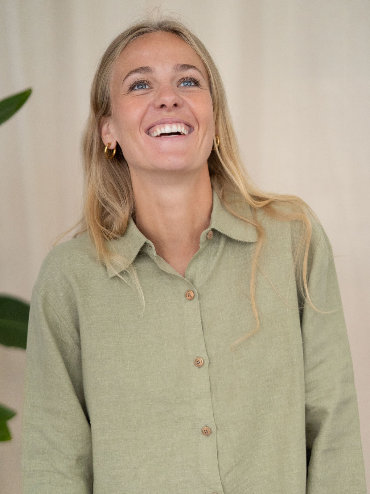 Eine junge, blonde Frau lächelt strahlend in die Kamera und trägt eine Frauen Langarm-Leinenbluse aus 100% Leinen. Made in Portugal, fair gefertigt in der Farbe salbei.
