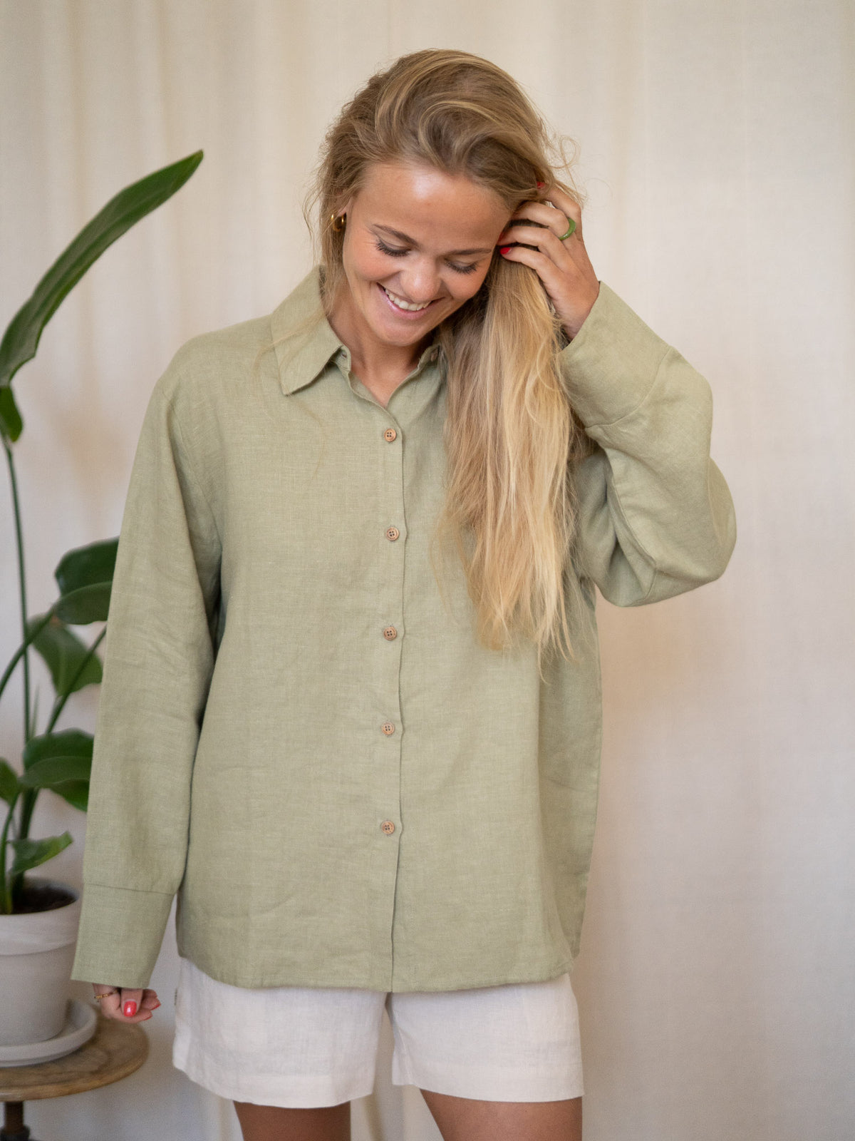 Eine junge, blonde Frau lächelt und blickt dabei nach unten. Sie trägt eine Frauen Langarm-Leinenbluse aus 100% Leinen. Made in Portugal, fair gefertigt in der Farbe salbei.