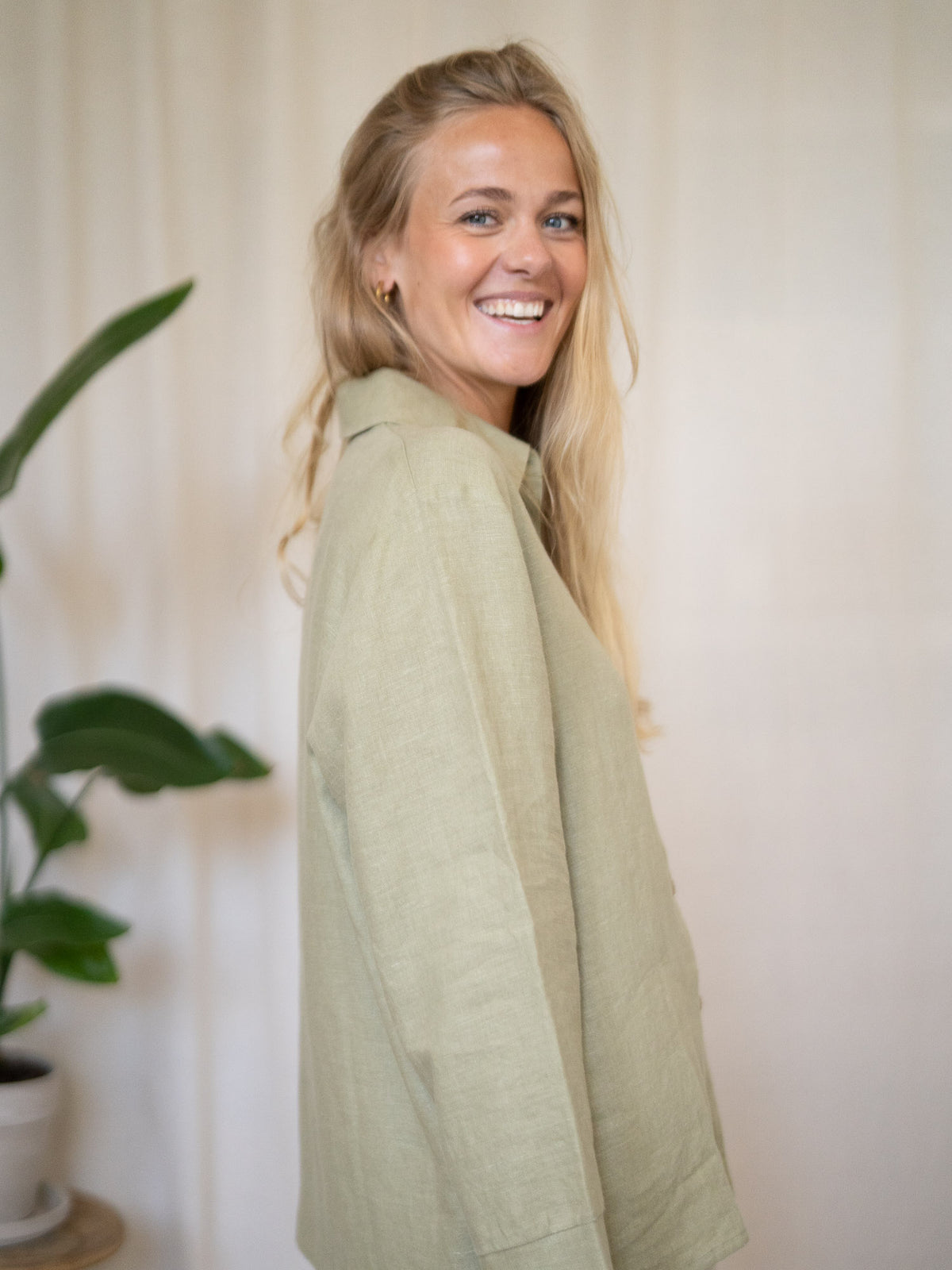 Eine junge, blonde Frau lächelt und steht seitlich zur Kamera. Dabei trägt sie eine Frauen Langarm-Leinenbluse aus 100% Leinen. Made in Portugal, fair gefertigt in der Farbe salbei.