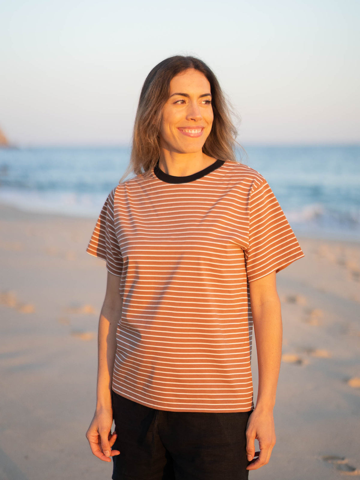 Gestreiftes Fair Fashion Frauen T-Shirt in karamell, schwarz und weiss von FUXBAU aus Biobaumwolle. Gefertigt unter GOTS-Bedingungen in Portugal und getragen am Strand in Portugal. Getragen von einer jungen Frau am Strand in Südportugal.