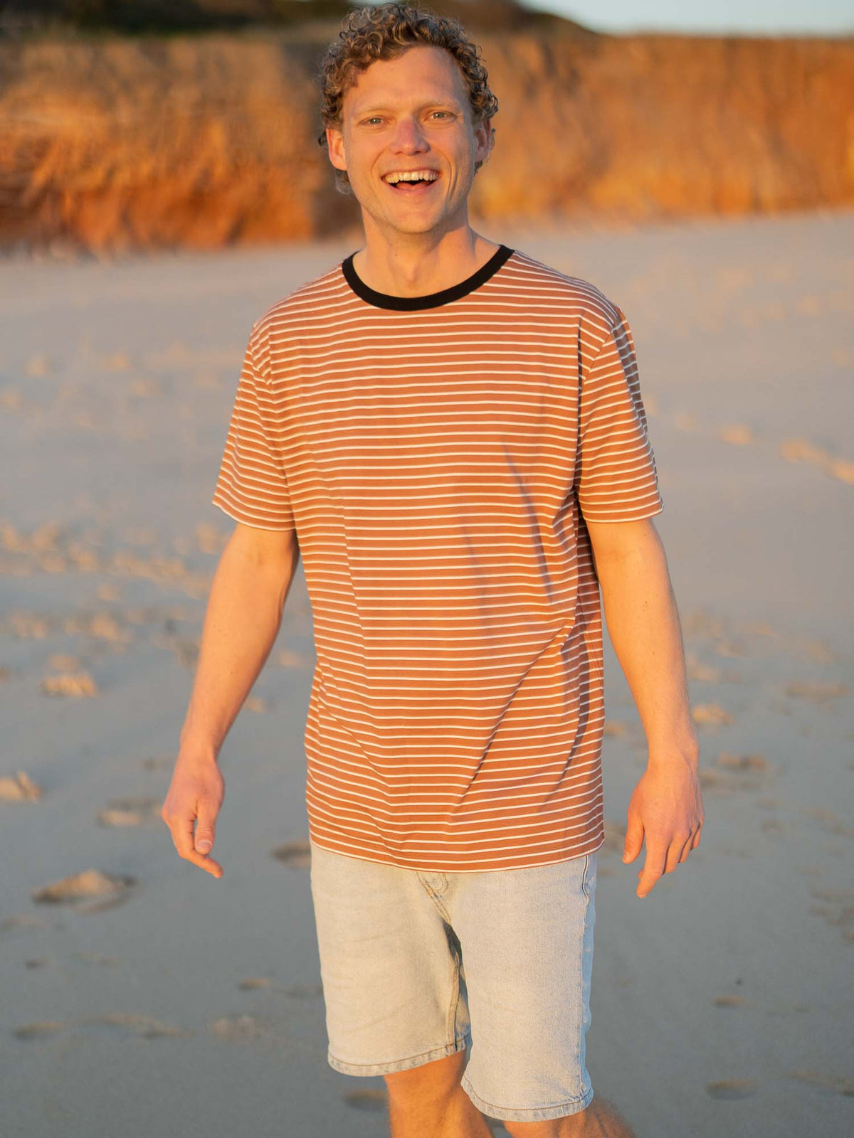 Gestreiftes Fair Fashion T-Shirt in karamell, schwarz und weiss von FUXBAU aus Biobaumwolle. Gefertigt unter GOTS-Bedingungen in Portugal und getragen am Strand in Portugal. Getragen von einem jungen blondem Mann in Portugal.