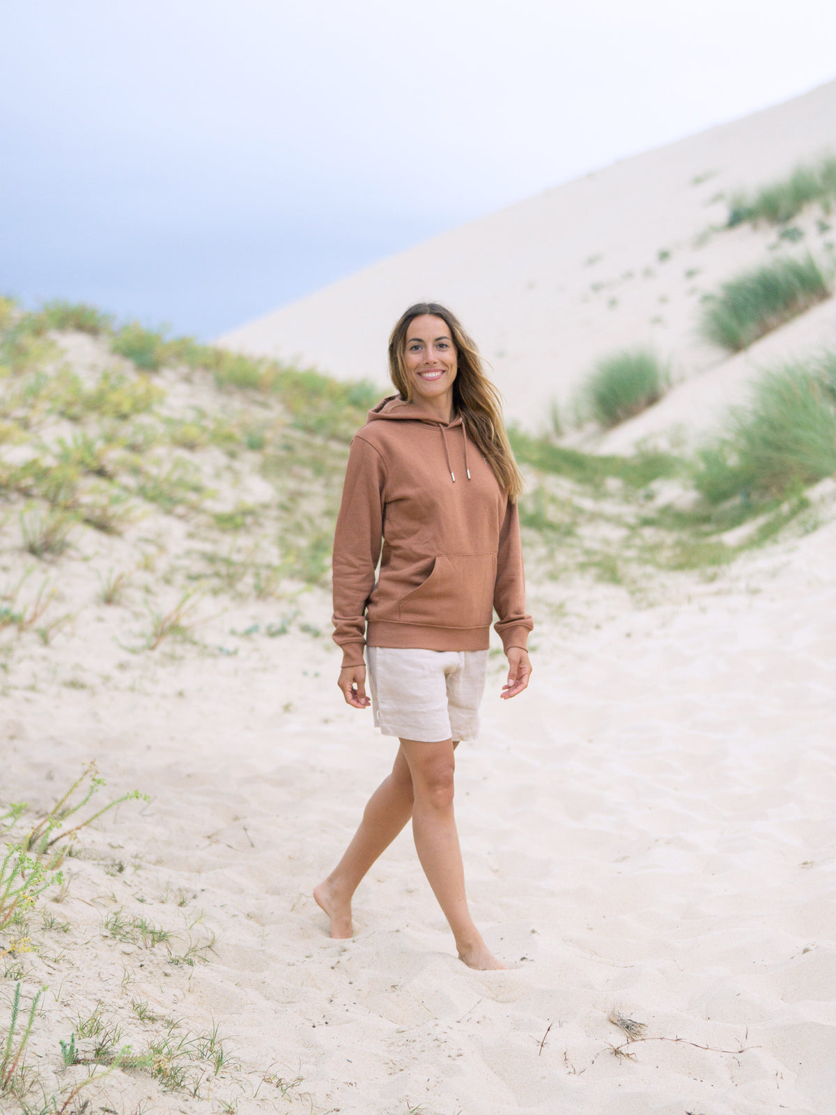 Unsere Freundin Rachel trÀgt unseren unisex Fair Fashion Hoodie in karamell am Strand aus 100% GOTS zertifizierter Biobaumwolle und made in Portugal