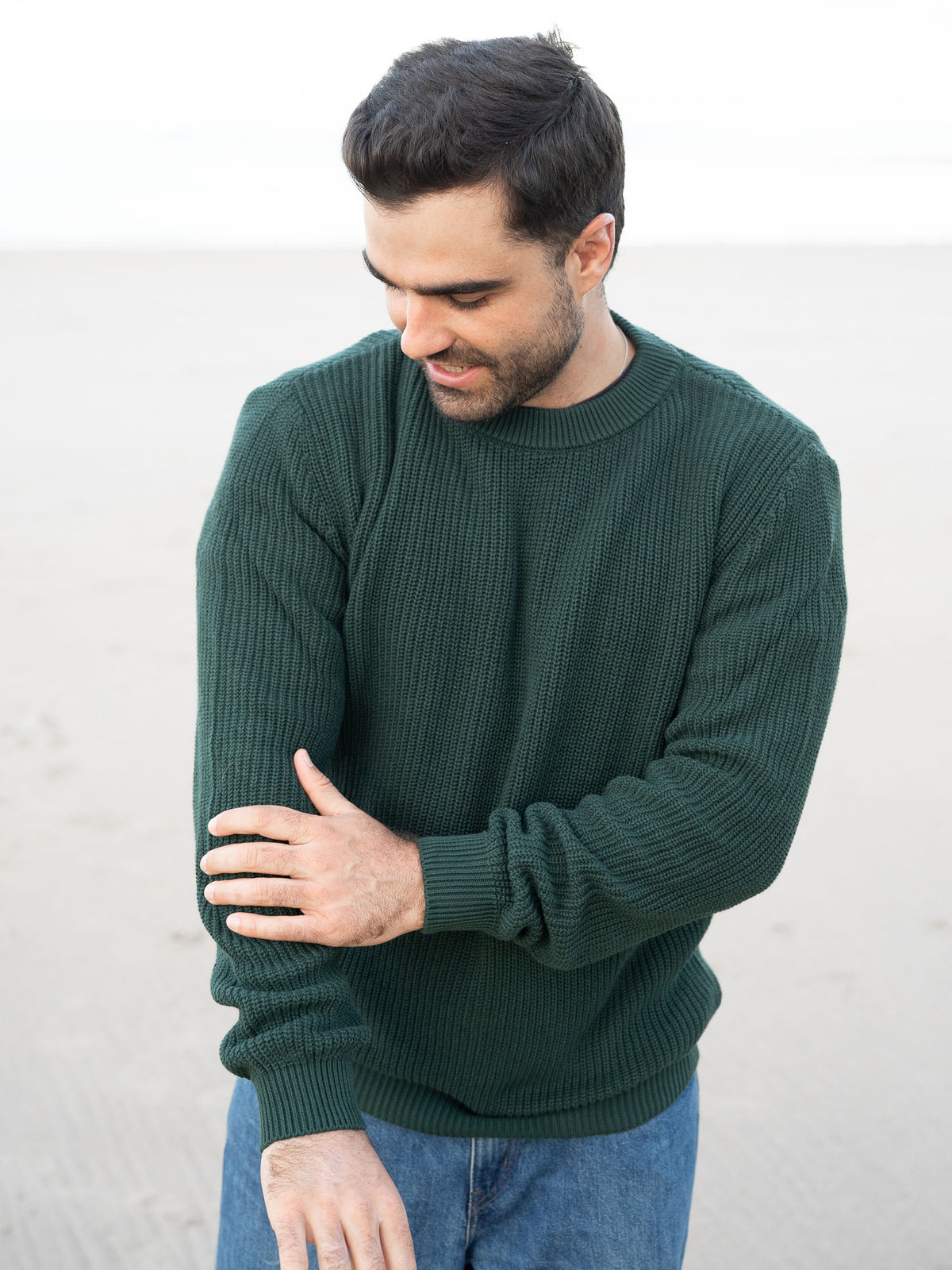 Junger, schwarzhaariger Mann trägt einen Fair Fashion Strickpullover von FUXBAU in grün aus GOTS-zertifizierter Biobaumwolle am Strand in Spanien.