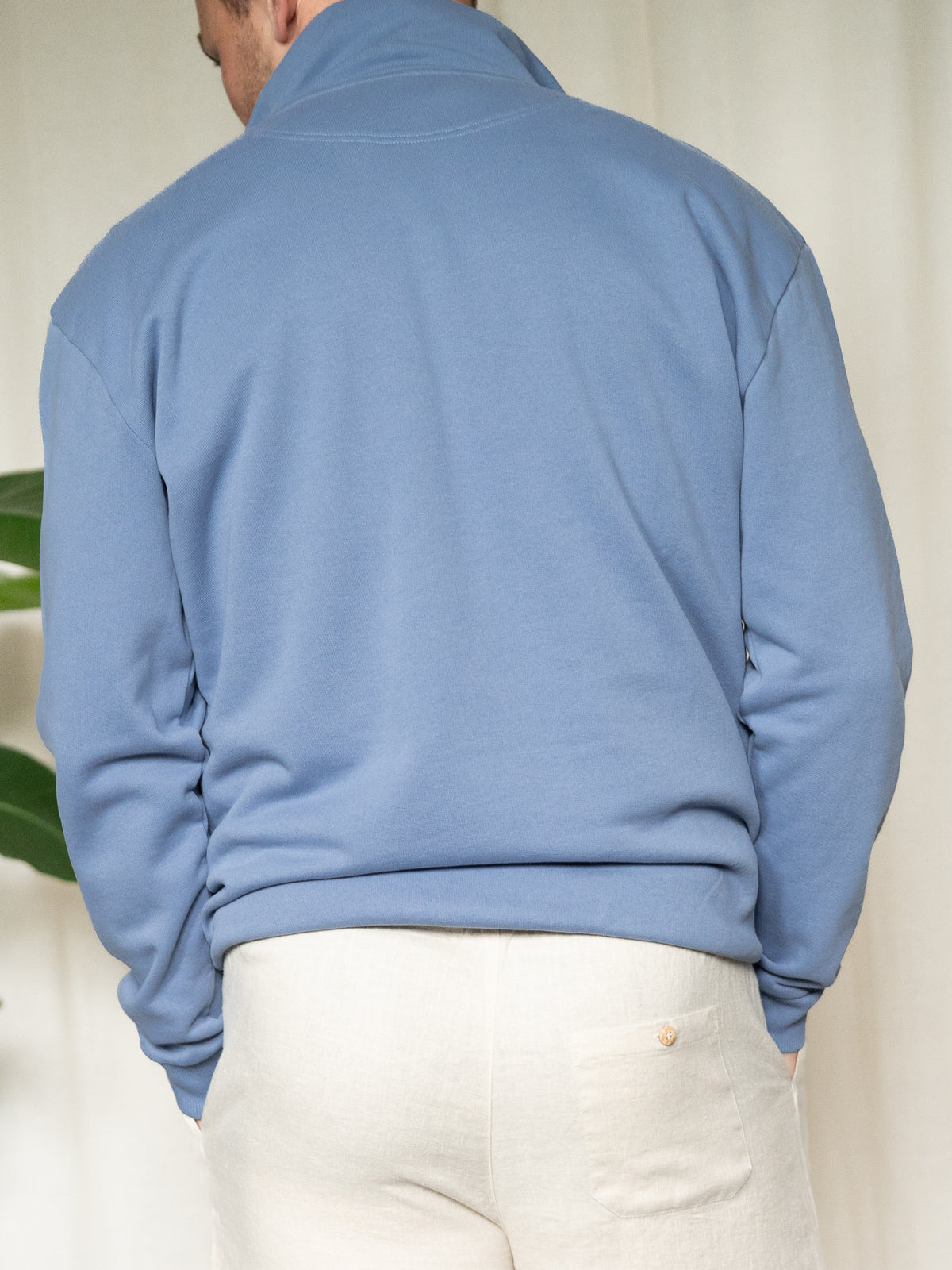 Rückansicht eines Mannes im himmelblauen FUXBAU Half-Zip Sweater, kombiniert mit heller Leinenhose.