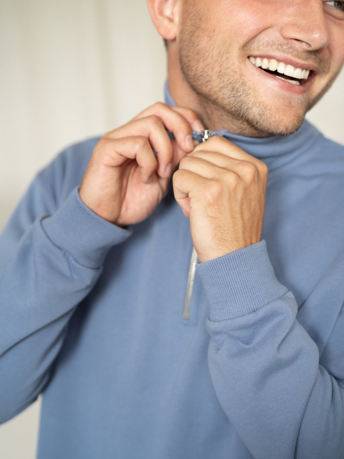 Mann schließt lächelnd den Reißverschluss des himmelblauen Half-Zip Sweaters.