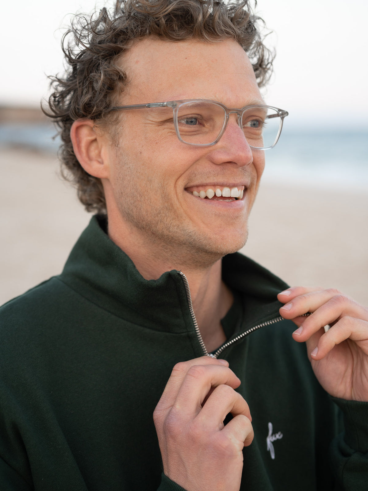 Junger, blonder Mann trägt einen Fair Fashion Männer Half Zip Sweater von FUXBAU in grün aus gots zertifizierter Biobaumwolle Made in Portugal mit YKK Reisverschlüssen am Strand in Portugal.