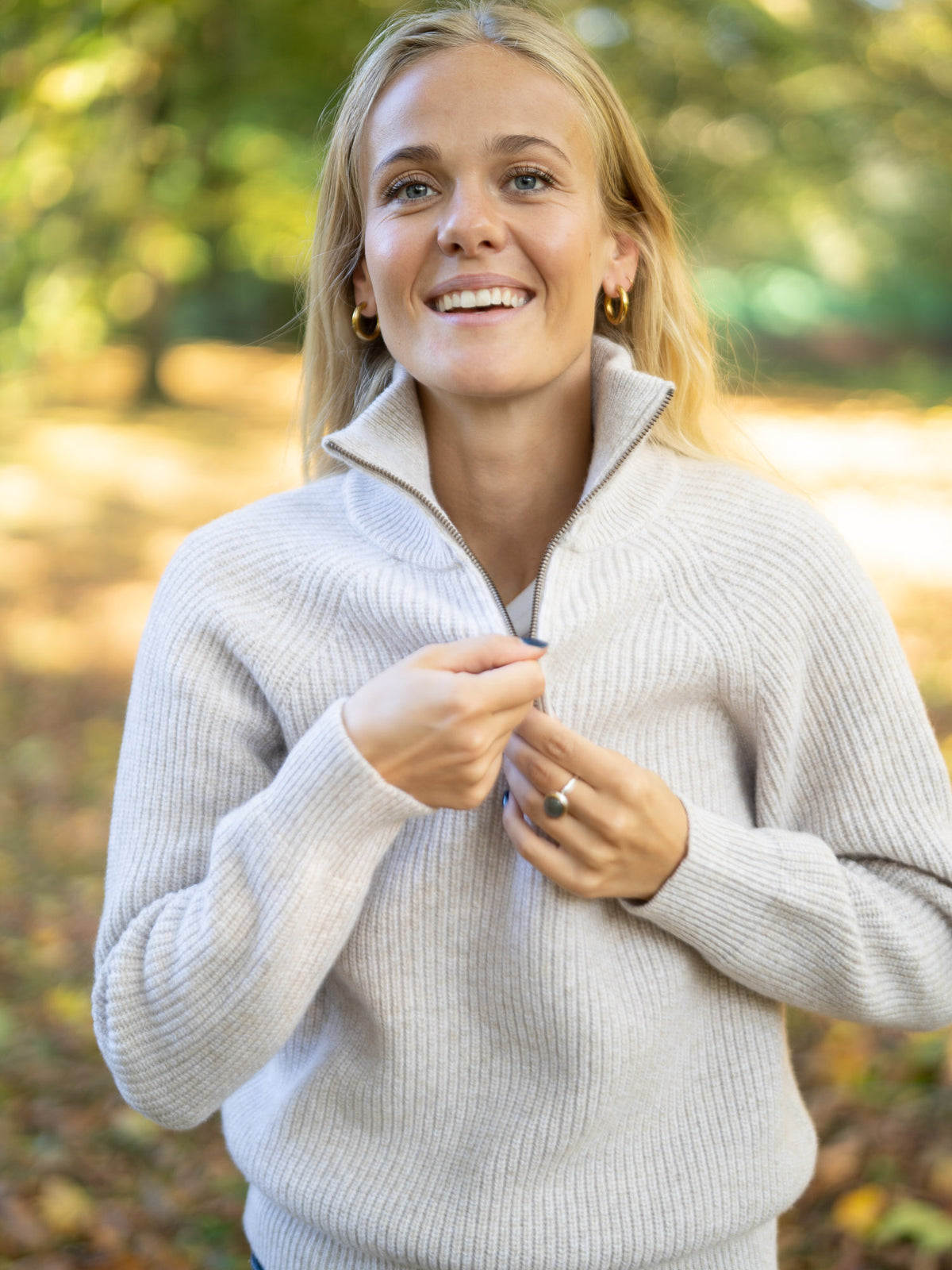 Eine junge, blonde Frau trägt einen fair produzierten Fair Fashion Troyer von FUXBAU aus 100% Merinowolle in der Natur im Herbst.