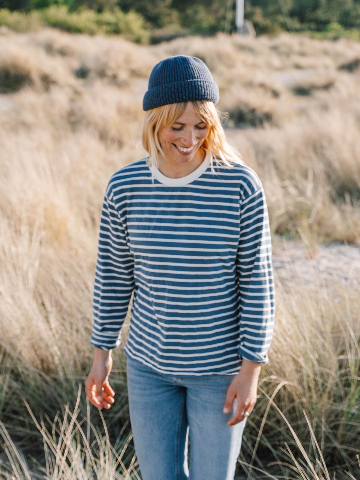 Junge, blonde Frau trĂ€gt ein maritimes FUXBAU Fair Fashion Outfit am Strand bestehend aus einem gestreiftem Longsleeve in creme blau, jeans und fischermĂŒtze aus Bio-Baumwolle.