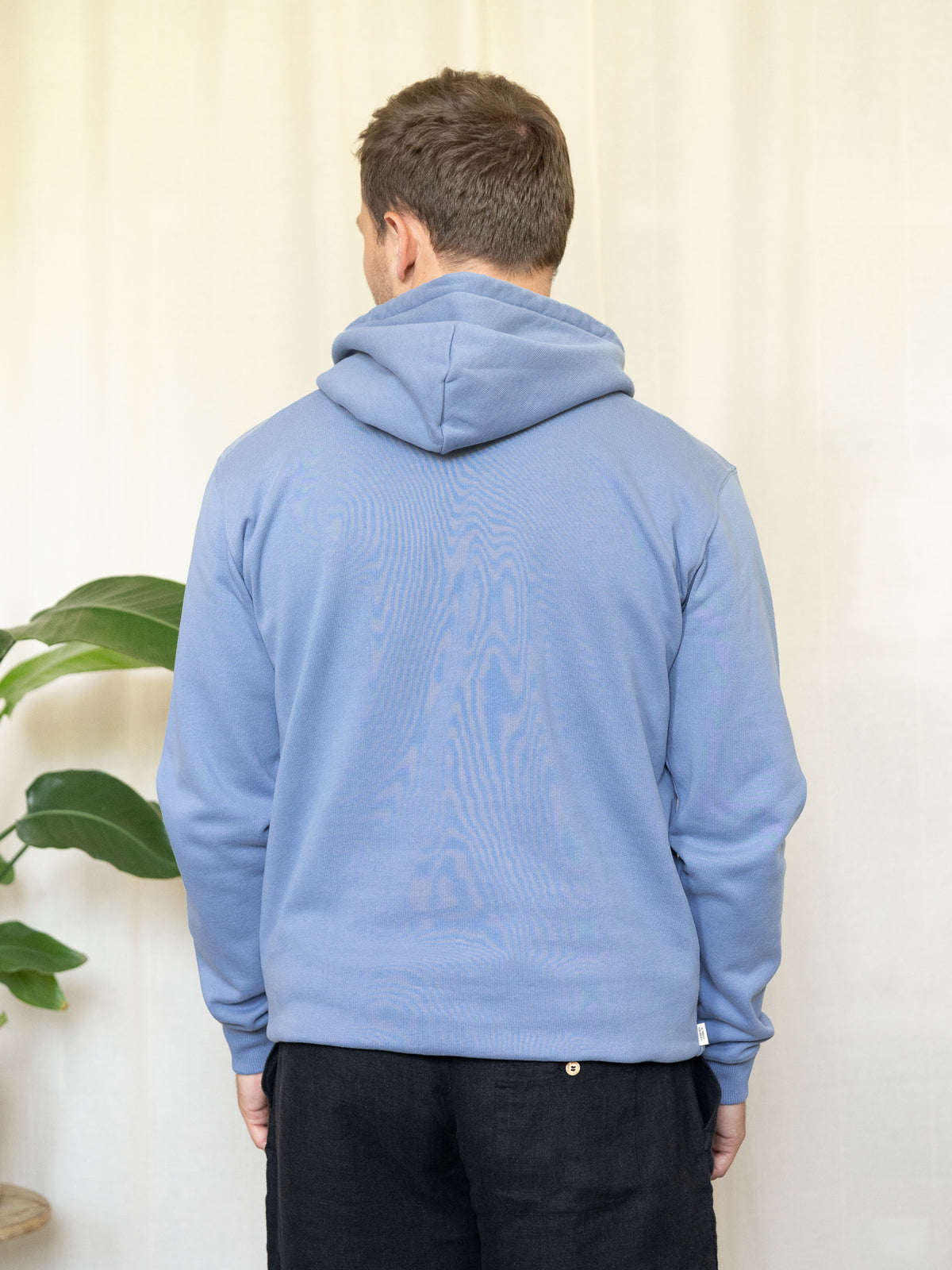 RĂŒckansicht eines himmelblauen Unisex-Hoodies aus Biobaumwolle, getragen von einer Person mit kurzen Haaren.