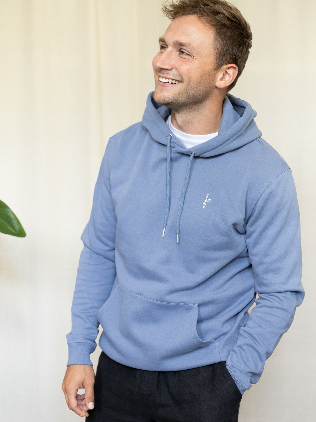Frontalansicht eines Mannes mit himmelblauem Hoodie aus Biobaumwolle und schwarzer Hose, lÀchelnd.