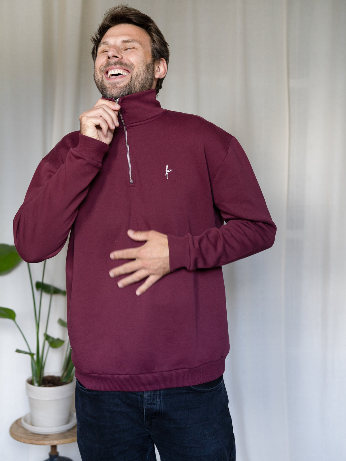 Mann zieht den Reißverschluss des bordeauxfarbenen FUXBAU Half-Zip Sweaters hoch – Pullover aus Biobaumwolle.