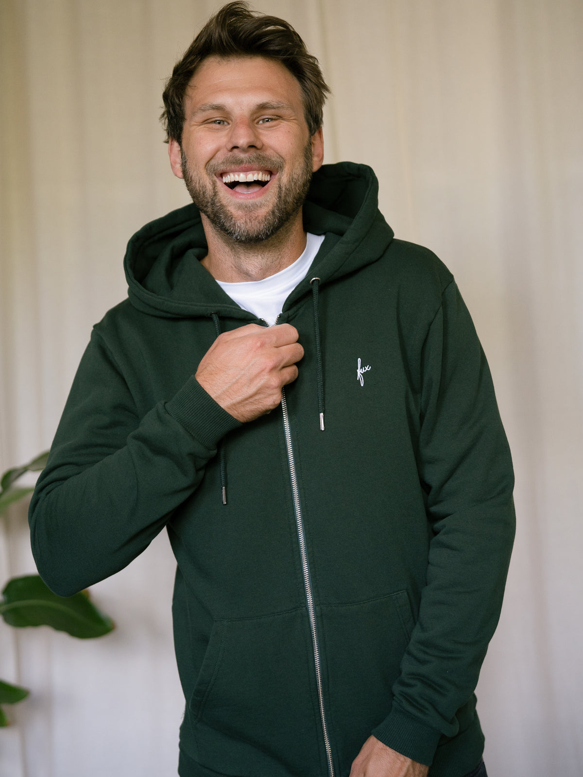 FUXBAU Sweatjacke Männer grün – Modell lacht mit geschlossener Jacke und Kapuze, fair in Portugal hergestellt aus 100% Biobaumwolle
