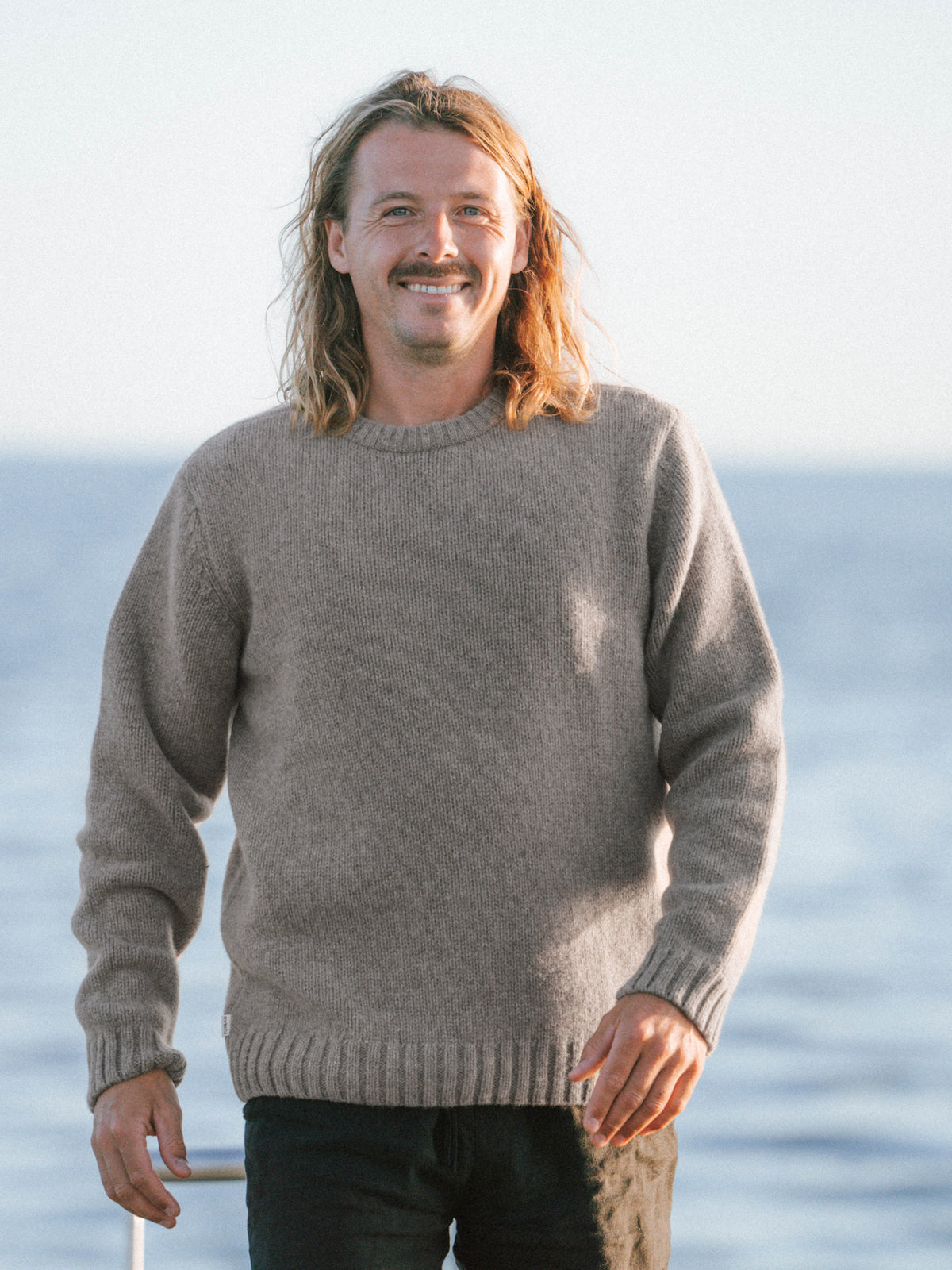 Ole trägt am Meer den Männer Merino Strickpullover in braunmeliert der in Deutschland hergestellt wird.