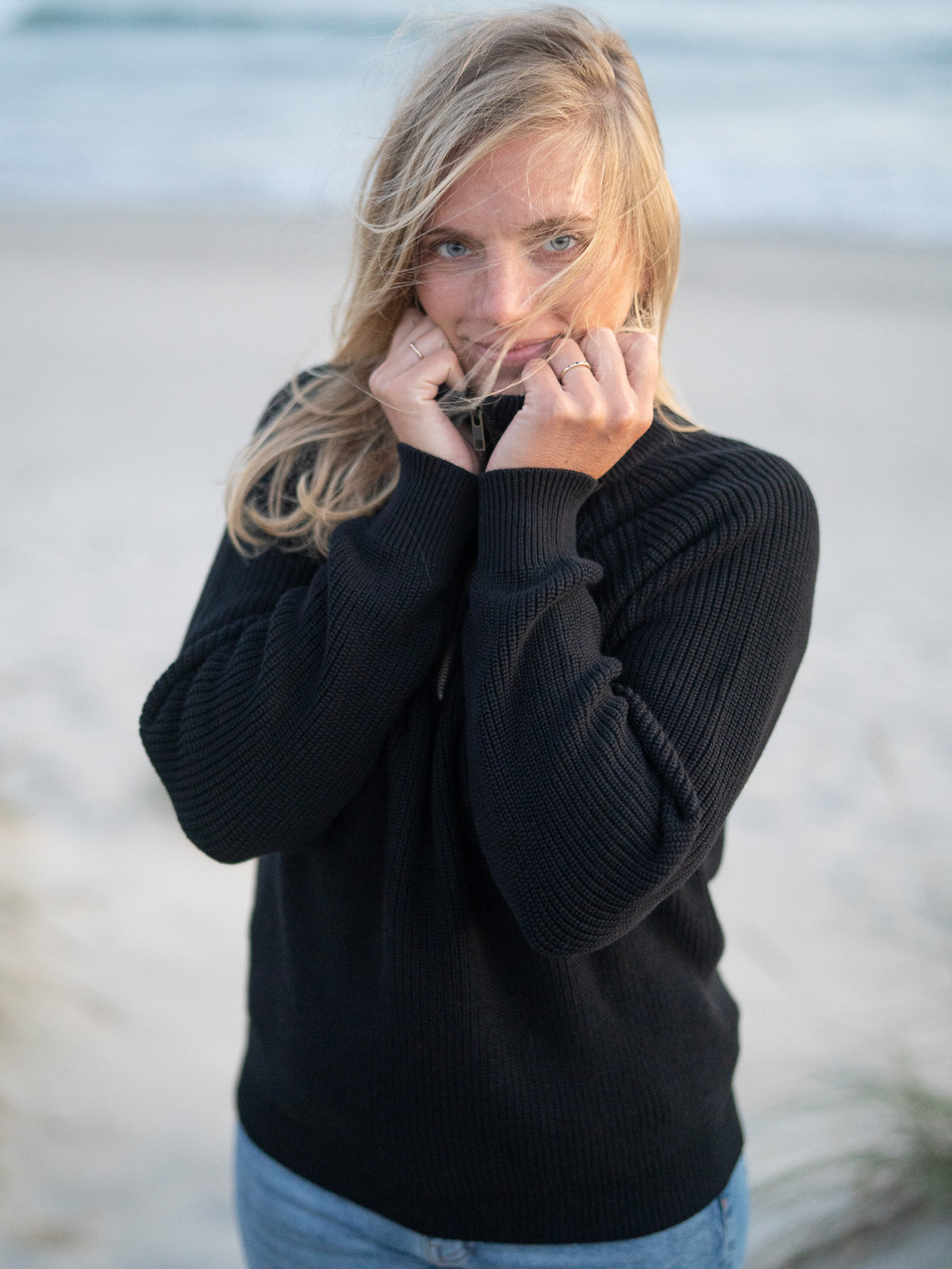 Eine junge, blonde Frau trägt einen Fair Fashion 4Seasons Troyer aus Biobaumwolle Made in Italy vom Fair Fashion Label FUXBAU am Strand in Portugal. Frontalansicht.
