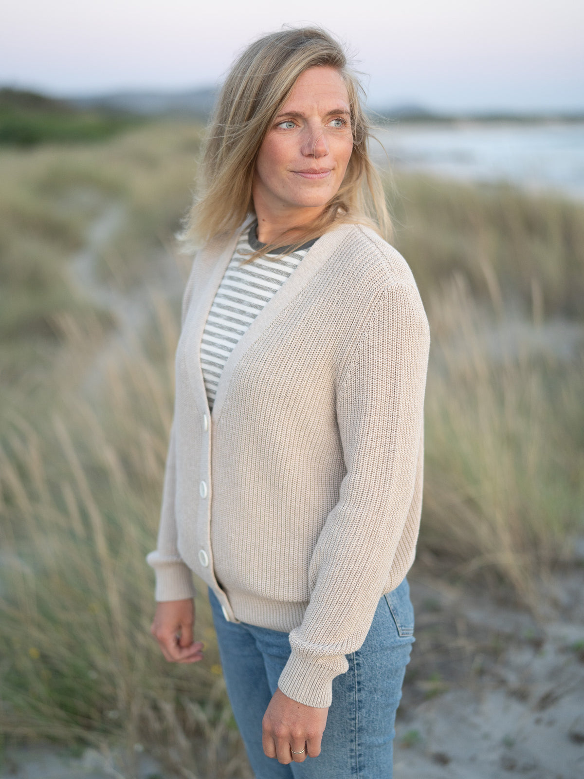 Eine junge, blonde Frau trägt eine beige melierte 4Seasons Strickjacke vom Fair Fashion Modelabel FUXBAU aus 100% GOTS Biobaumwolle am Strand in Portugal. Seitliche Ansicht.