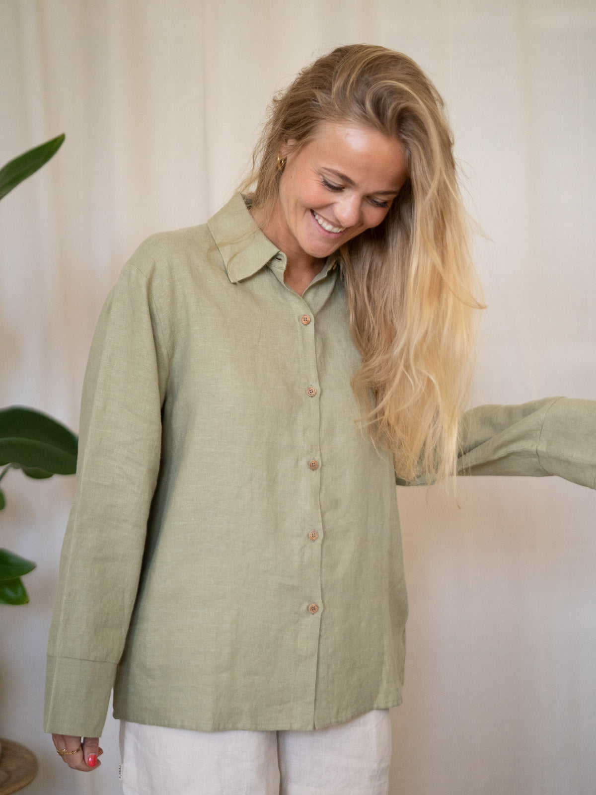 Eine junge, blonde Frau trägt eine Frauen Langarm-Leinenbluse aus 100% Leinen vom Fair Fashion Modelabel FUXBAU und lacht dabei. Gefertigt in Portugal unter fairen Bedingungen in der Farbe salbei.