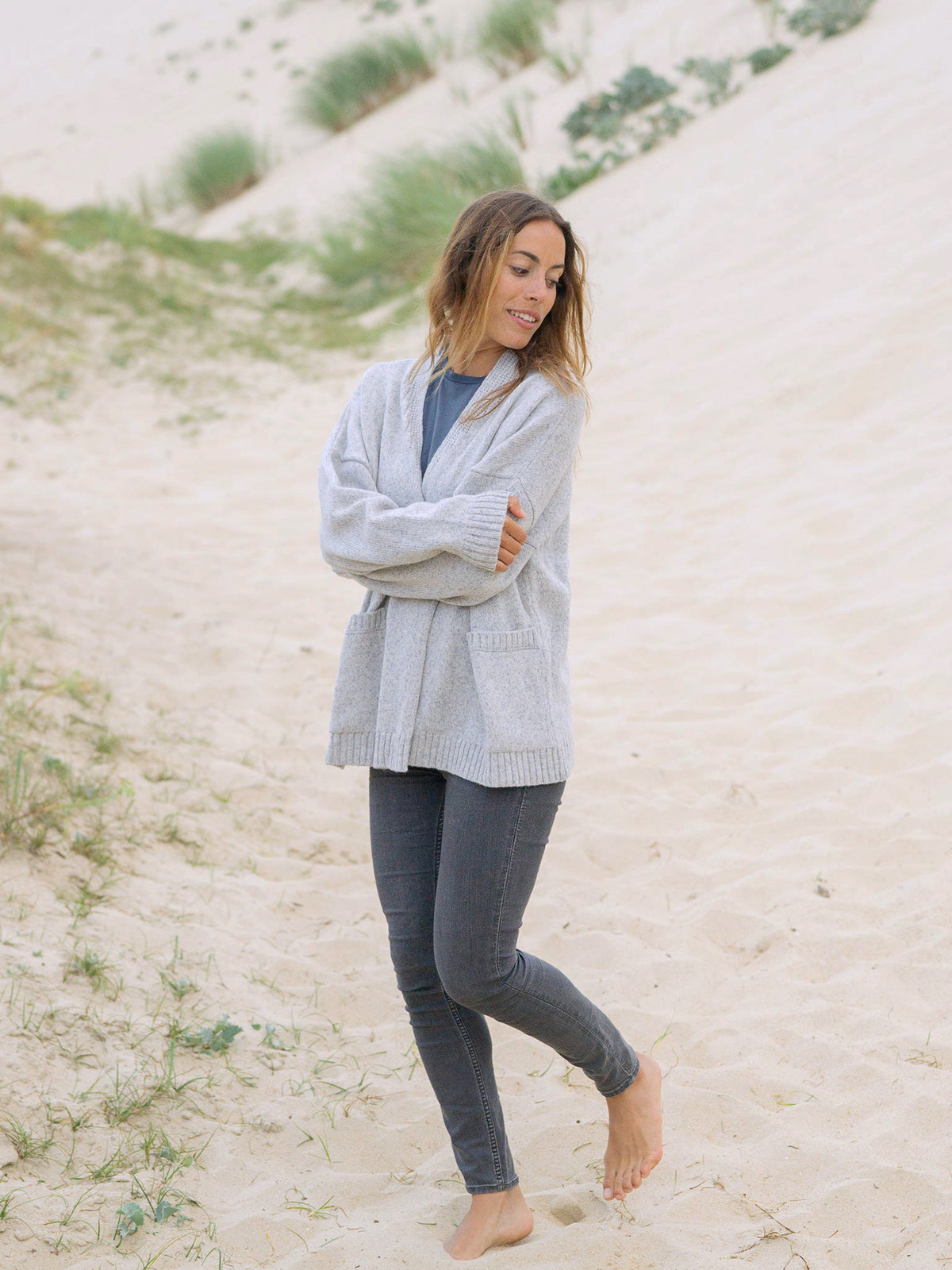 Graumelierter Oversized Frauen Cardigan aus 100% Merino Schurwolle von FUXBAU. Getragen von einem braunhaarigem Model am Strand in Fankreich.