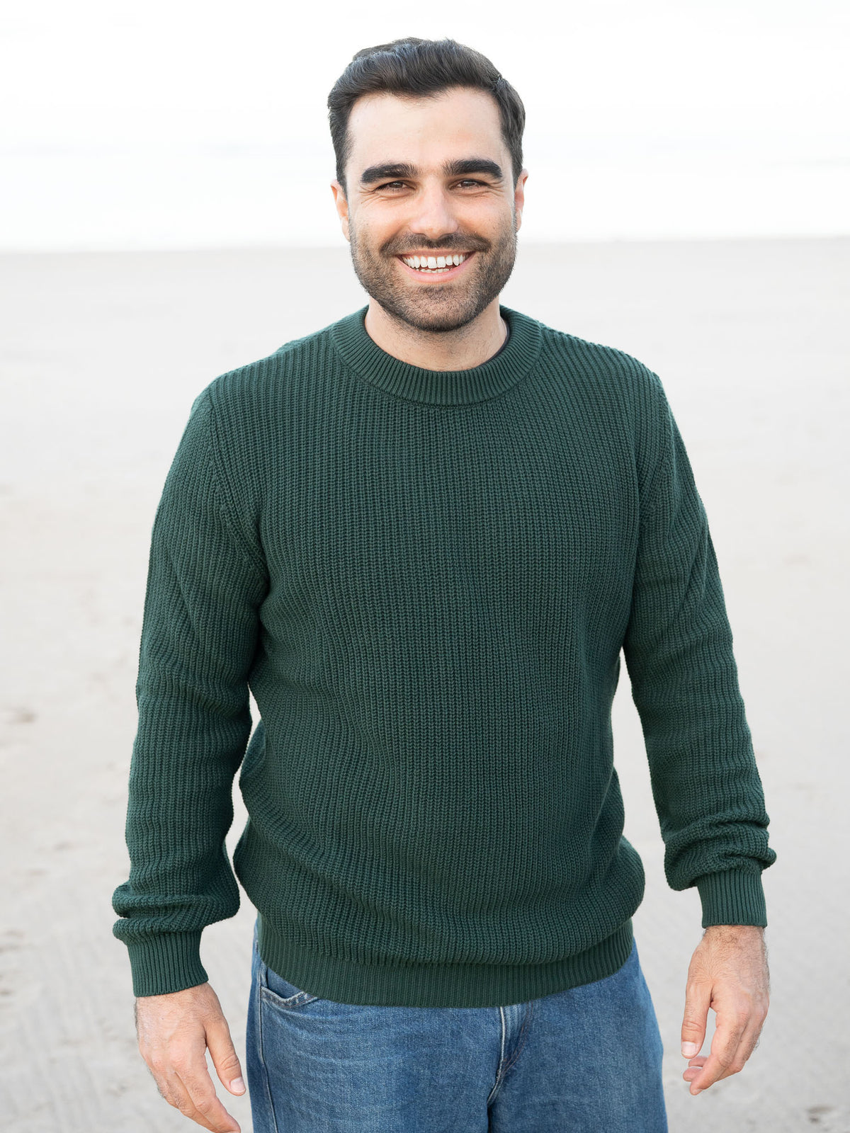 Junger, schwarzhaariger Mann trägt einen Fair Fashion Strickpullover von FUXBAU in grün aus GOTS-zertifizierter Biobaumwolle am Strand in Spanien.