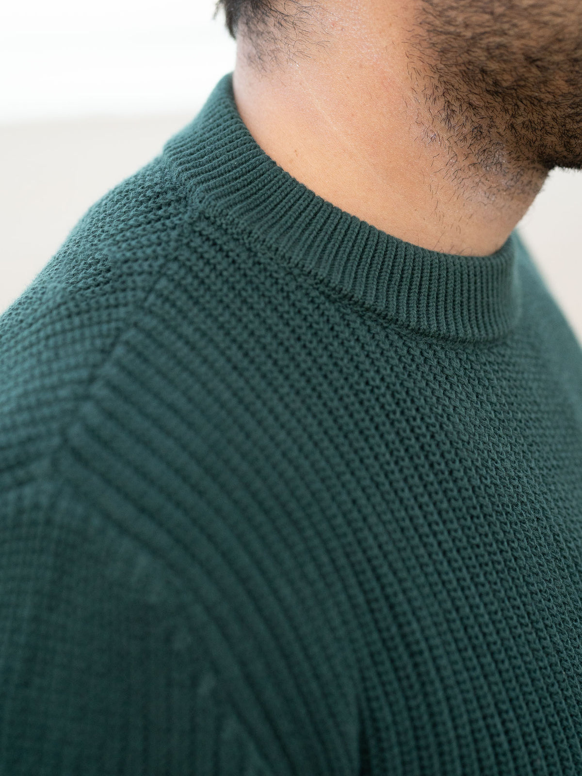 Junger, schwarzhaariger Mann trägt einen Fair Fashion Strickpullover von FUXBAU in grün aus GOTS-zertifizierter Biobaumwolle am Strand in Spanien.