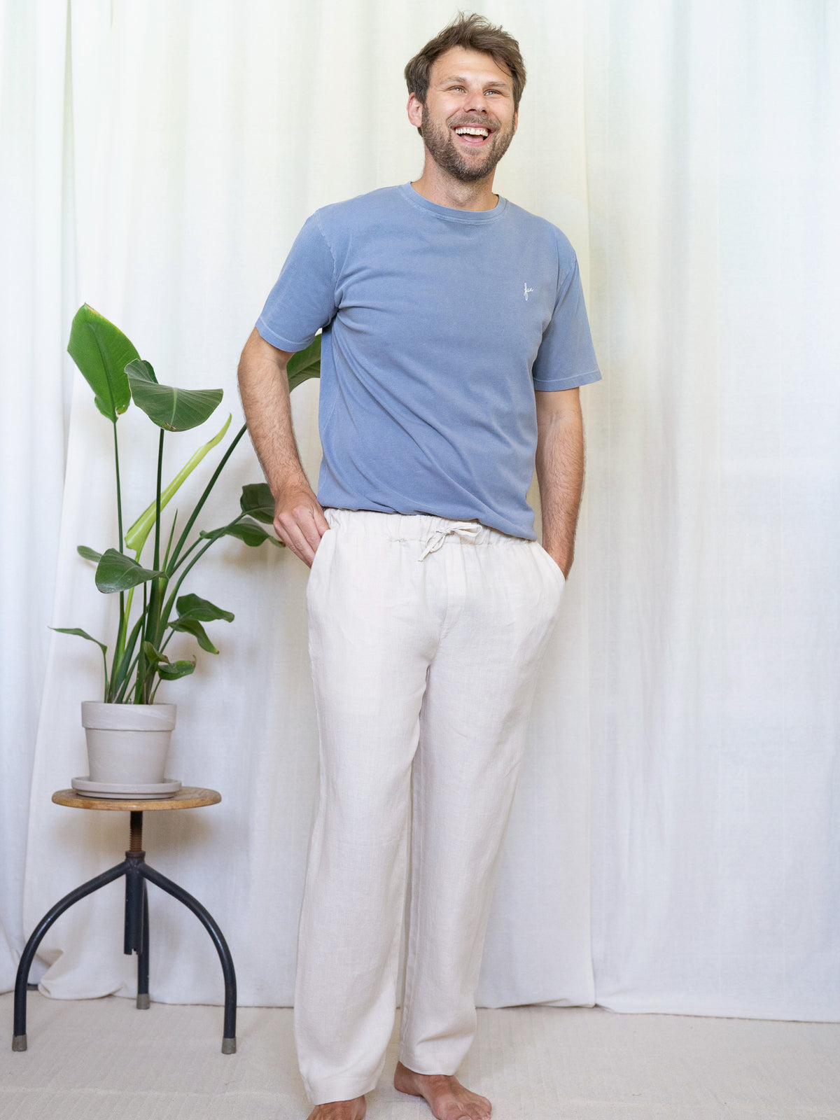 Ein junger Mann trägt eine Fair Fashion Leinenhose von FUXBAU in cremeweiss in Kombination mit einem blauen T-Shirt aus Biobaumwolle vor einem Vorhang.