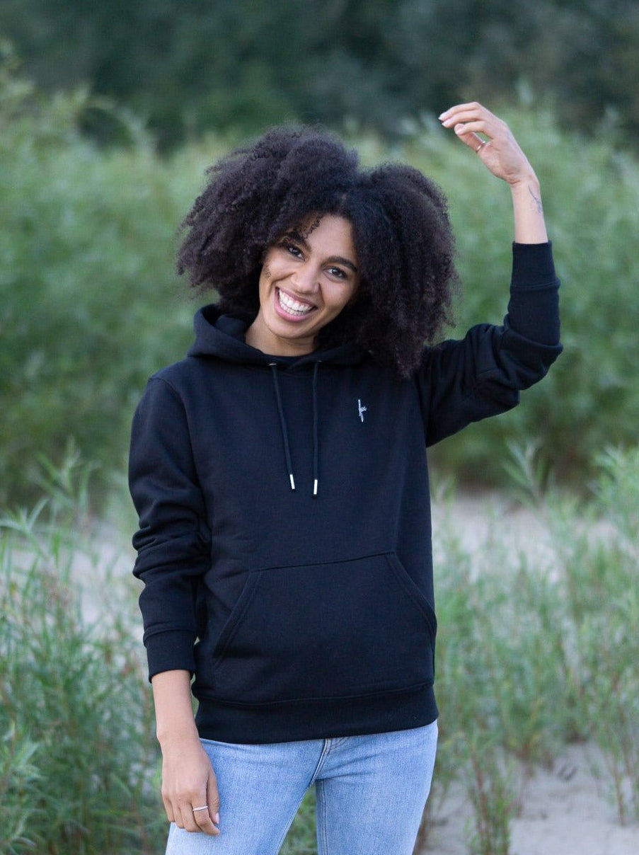 Junge, schwarzhaarige Frau trägt einen fairen Hoodie von FUXBAU in schwarz in Hamburg am Strand.