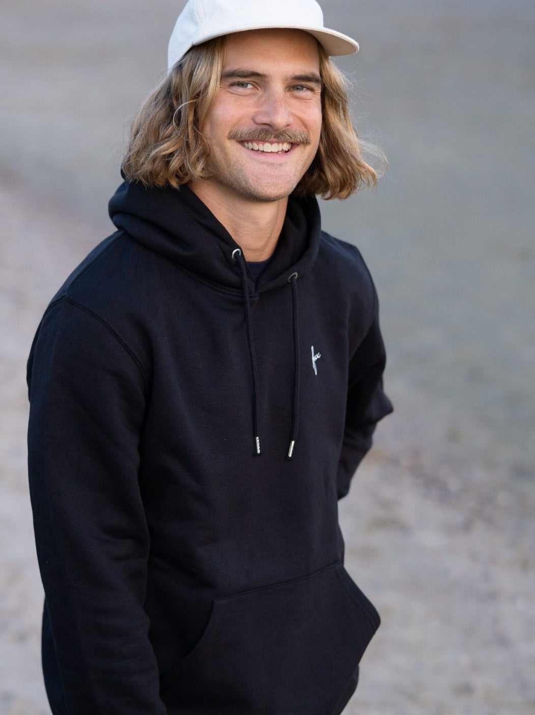 Junger, braunhaariger Mann trägt einen schwarzen FUXBAU Hoodie aus Biobaumwolle.